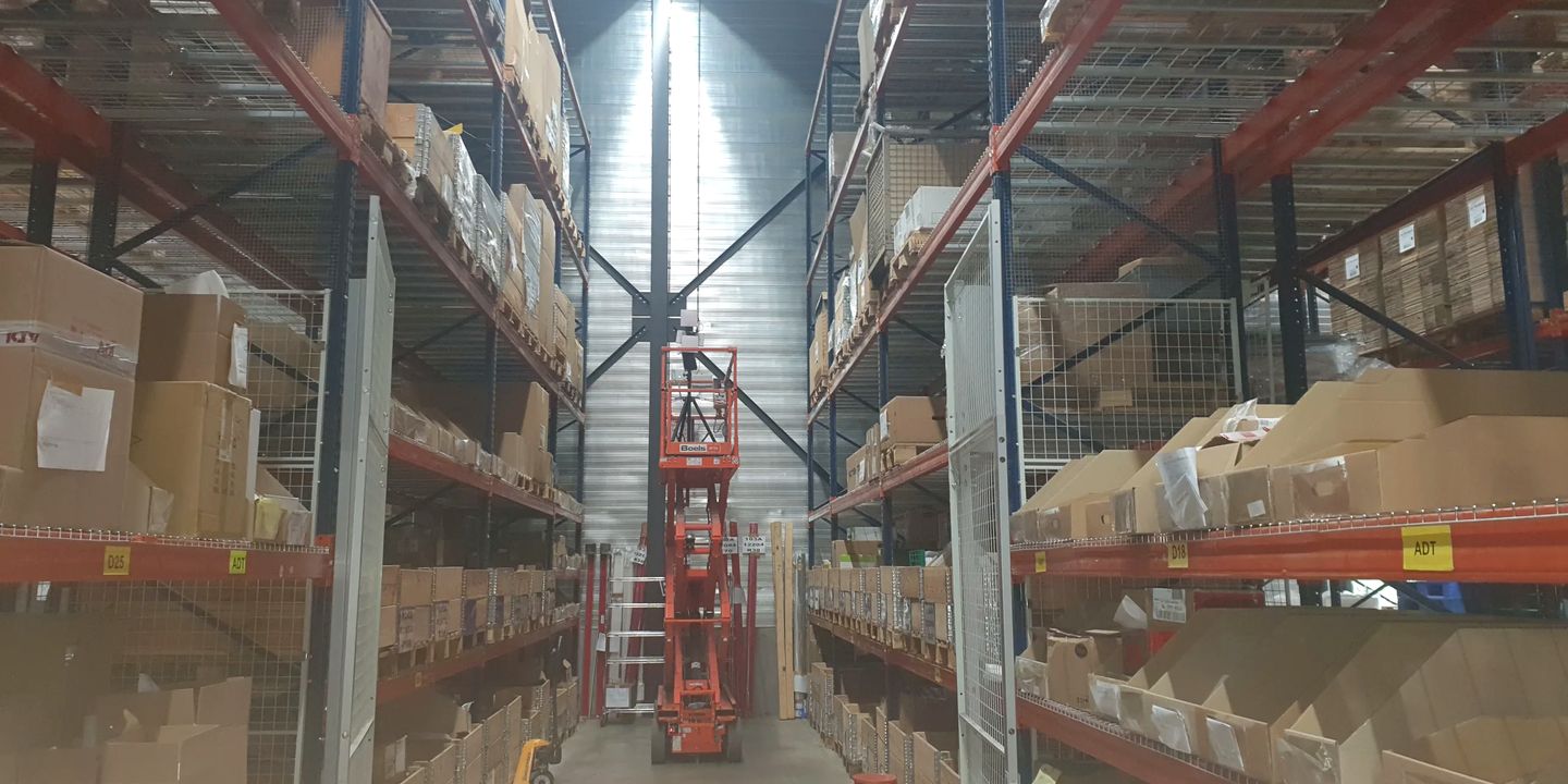 A Wi-Fi survey access point on a tripod mast, raised on an orange scissor lift in a narrow aisle between high-bay pallet racking.