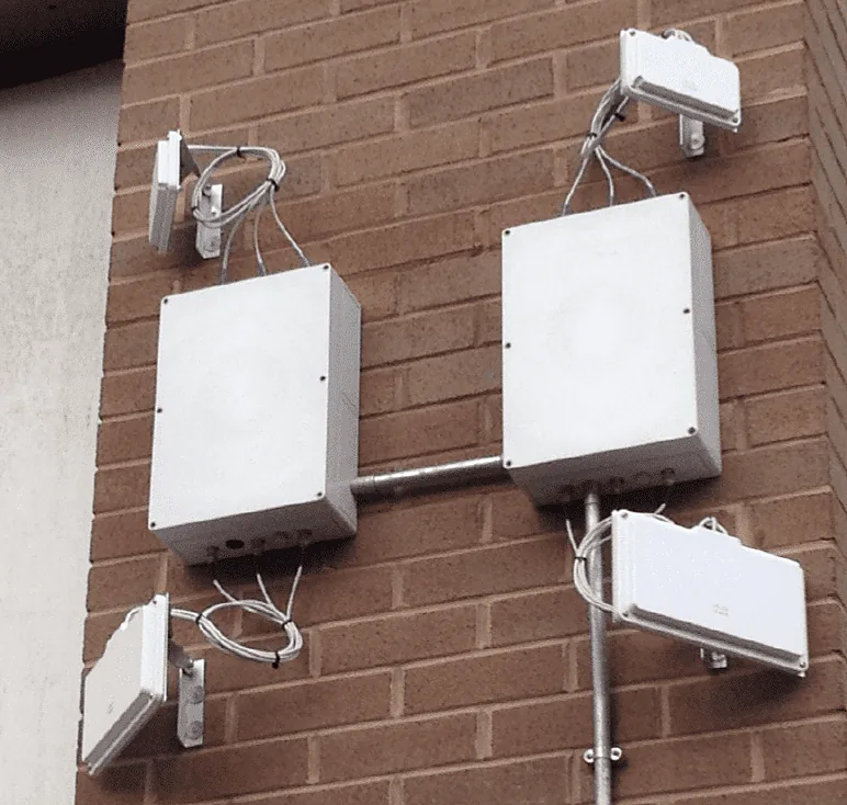 Two white DW WiFi outdoor enclosure kits fixed to a red brick wall and joined by galvanised conduit, with grey antenna leads running out to four external patch antennas on small brackets around them.