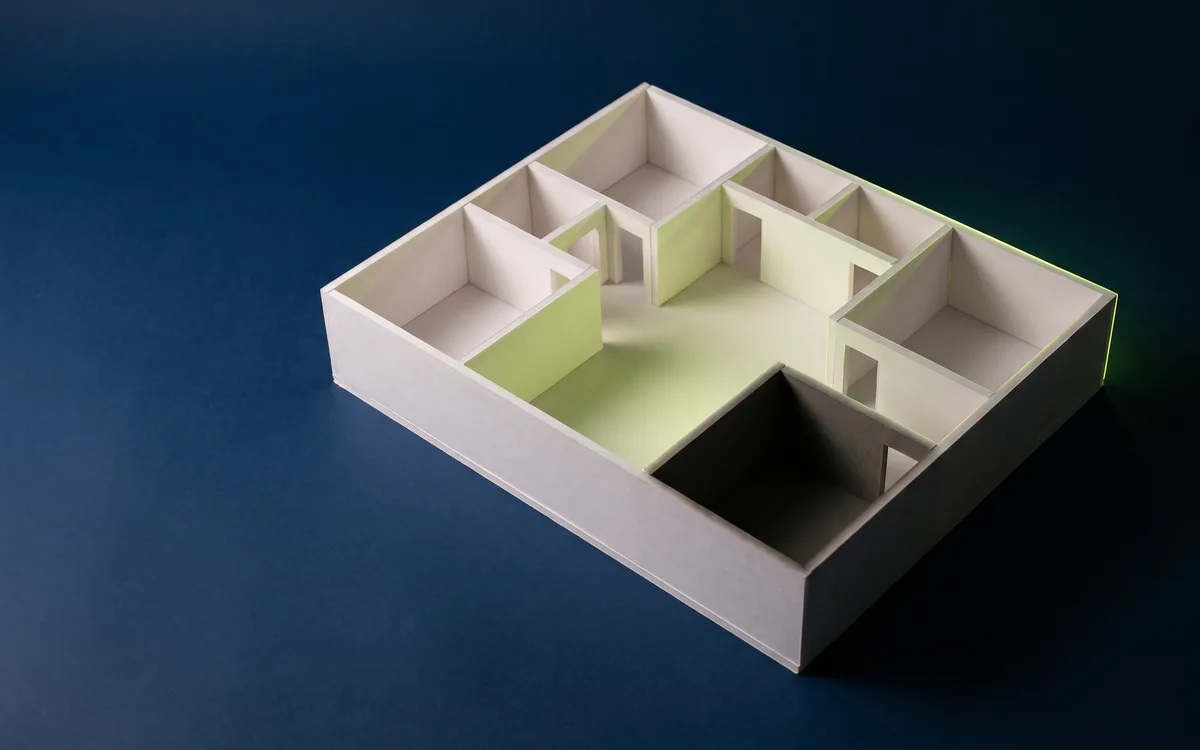 Illustration: a white foam-board model of a building floor with no roof, lime light pooling in the central rooms and one corner room left in shadow.