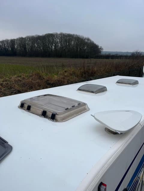 A cleaned caravan roof, white and even, with a rooflight, vent and satellite dish, ploughed fields and bare trees behind.