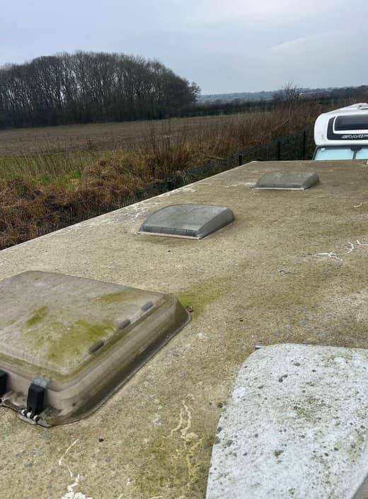 A caravan roof covered in green algae, the rooflight thick with growth, hedgerow and open fields behind.