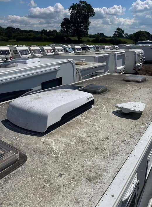 A caravan roof before cleaning, gritty and stained, with an air conditioning unit and vent fitted and a long row of stored caravans behind.