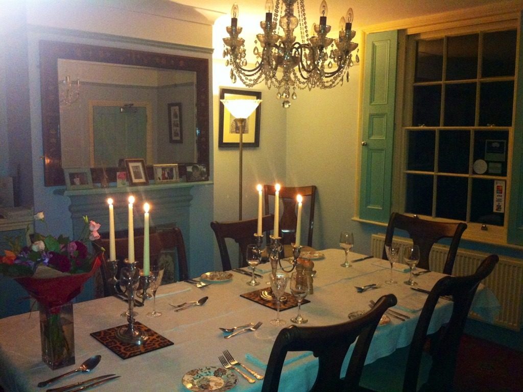 The same dining room at night, the long table dressed in white linen with a lit candelabra beneath a crystal chandelier