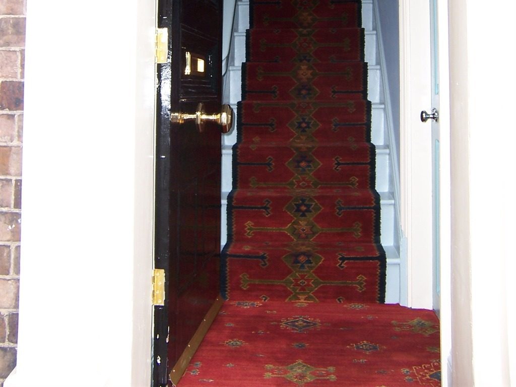 The open front door of the house with a brass knob and a red patterned runner climbing the staircase inside