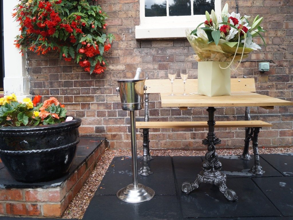 A bistro table on the brick patio set with champagne flutes, an ice bucket and flowers beneath a red begonia basket