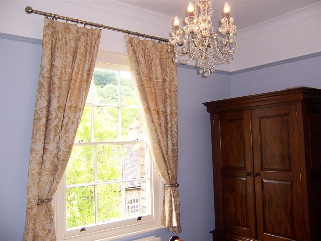 A pale-blue room with gold damask curtains tied back from a sash window that looks out over the wooded gorge, a crystal chandelier and a mahogany wardrobe