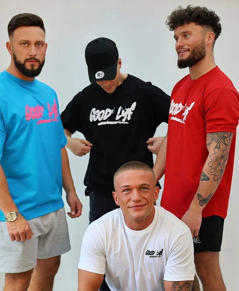 Four friends against a white studio wall in GoodLyf tees: oversized summer blue with the pink print, a black tee under a black cap, regular red, and white with the black wordmark.