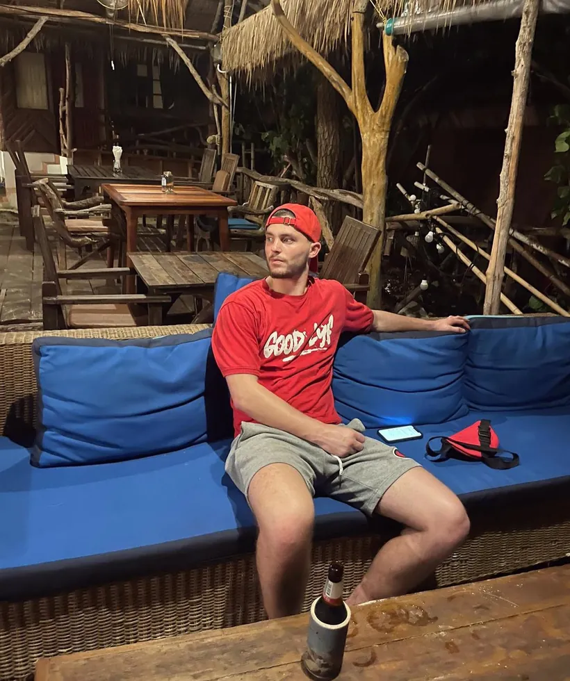 Red GoodLyf tee and a backwards red cap on holiday, stretched out on a rattan sofa at a beach bar.