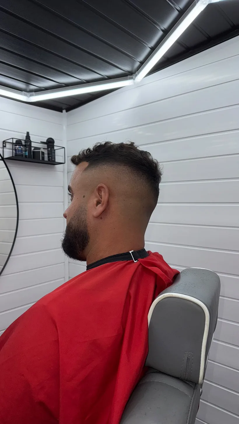 Back view of a high skin fade with textured curls left long on top, full beard, client in a red cape