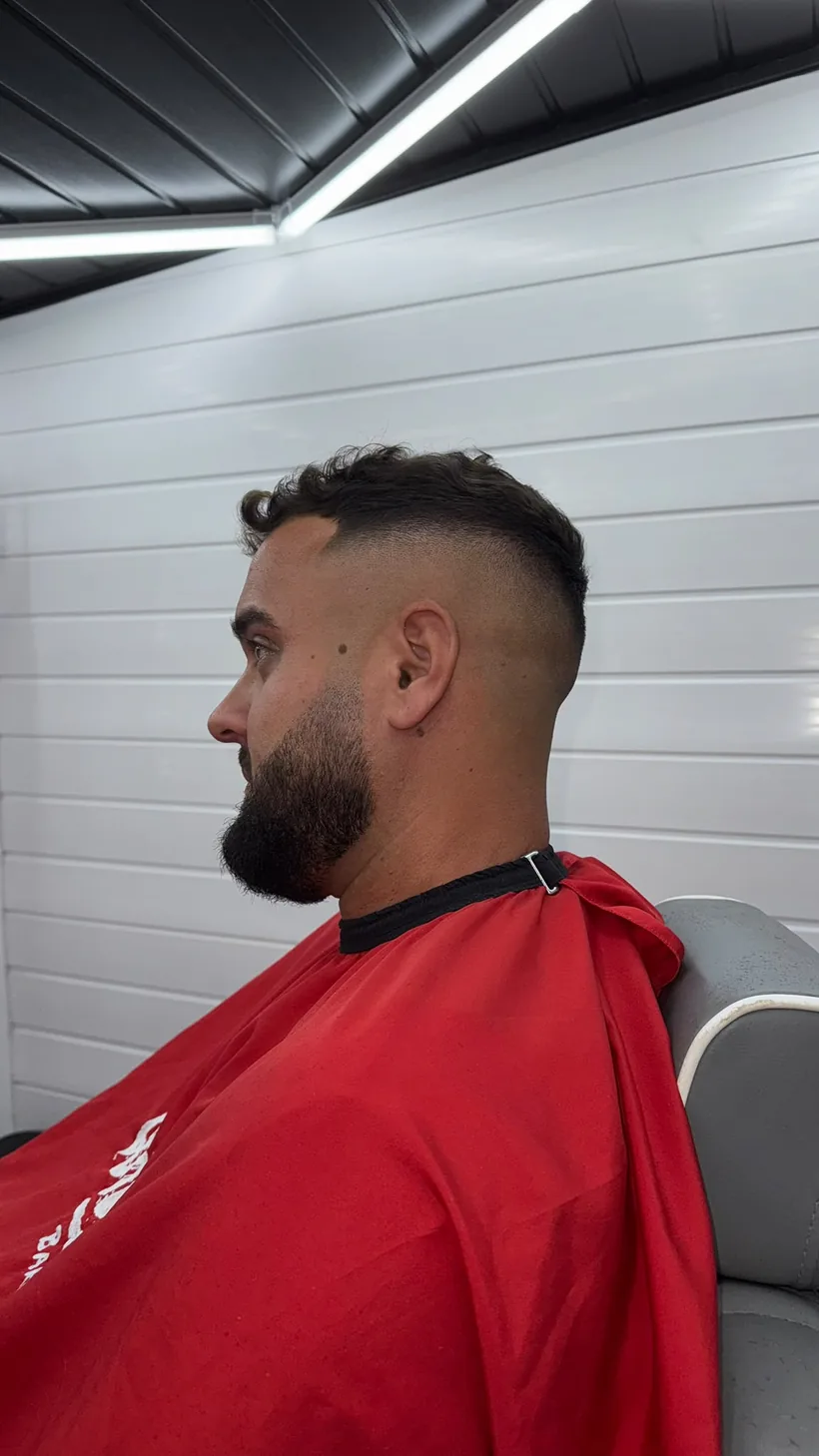 Side profile of the same high skin fade and lined-up full beard, client in a red cape