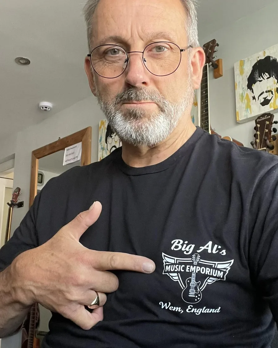 Jamie Davey, pointing at his Big Al's Music Emporium t-shirt, with acoustic guitars hanging on the wall behind him
