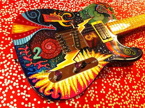 A hand painted psychedelic J. Davey Telecaster covered in sunburst rays, spirals and bright colour, on a red spotted backdrop