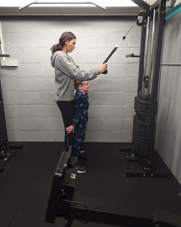 A young boy in a camouflage onesie standing at his mum's side while she trains on the cable machine at Let Luce.