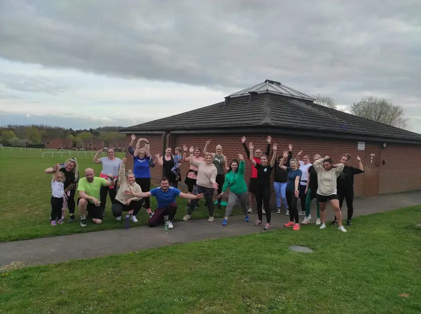 A big group of Lucy&rsquo;s clients, a child among them, on a grass field in front of a brick building, arms up and waving.