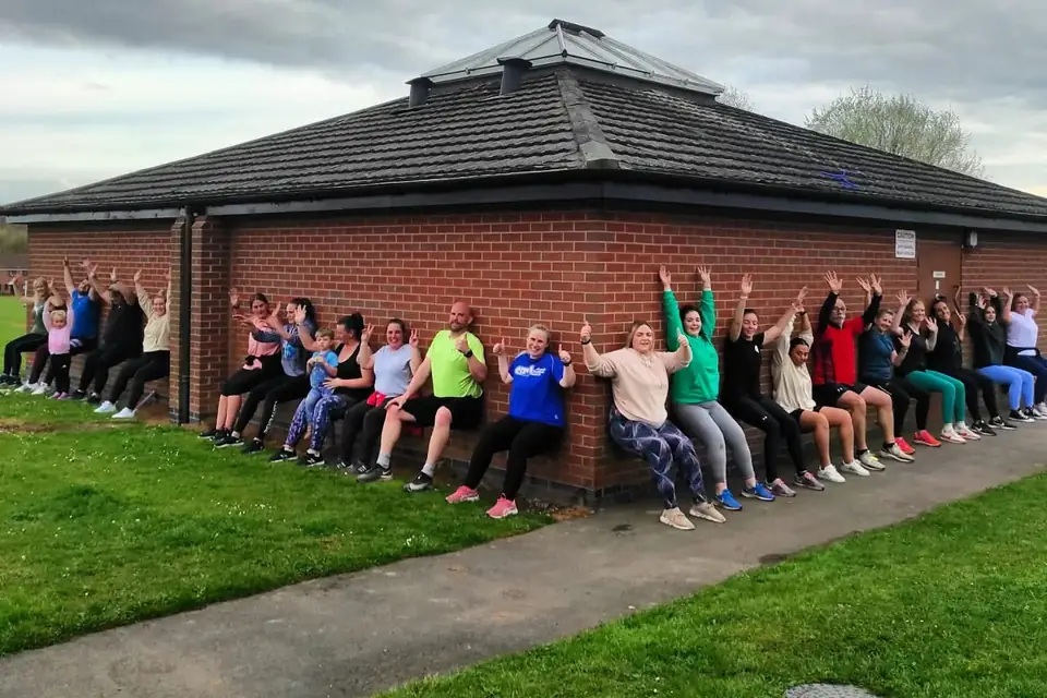 Lucy&rsquo;s clients doing a wall sit in a line against a brick building on the field, arms raised and thumbs up.