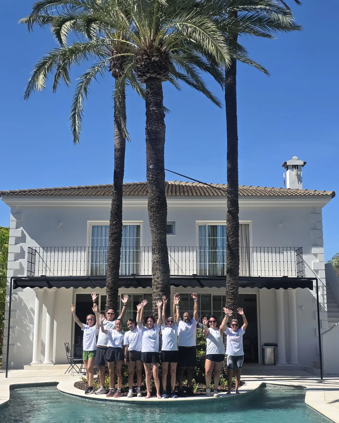 A retreat group cheering with arms up on the pool island in front of a white villa and palm trees.
