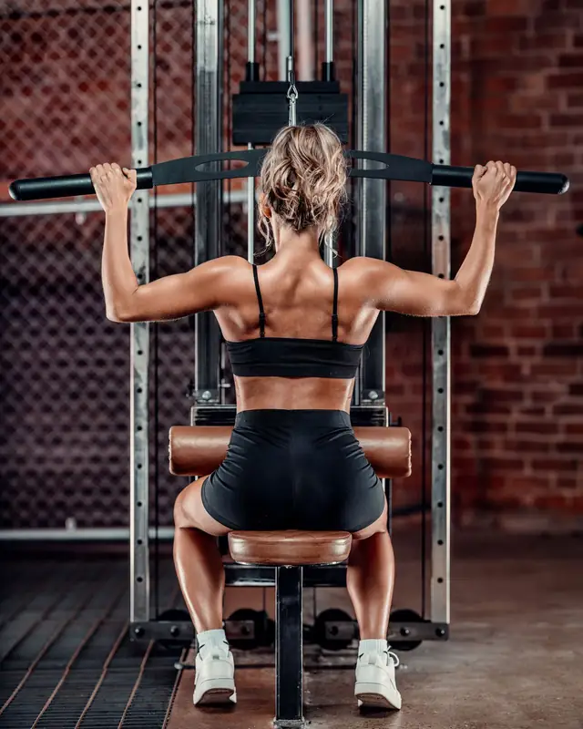 Lucy seen from behind doing a lat pulldown in her gym, against red brick