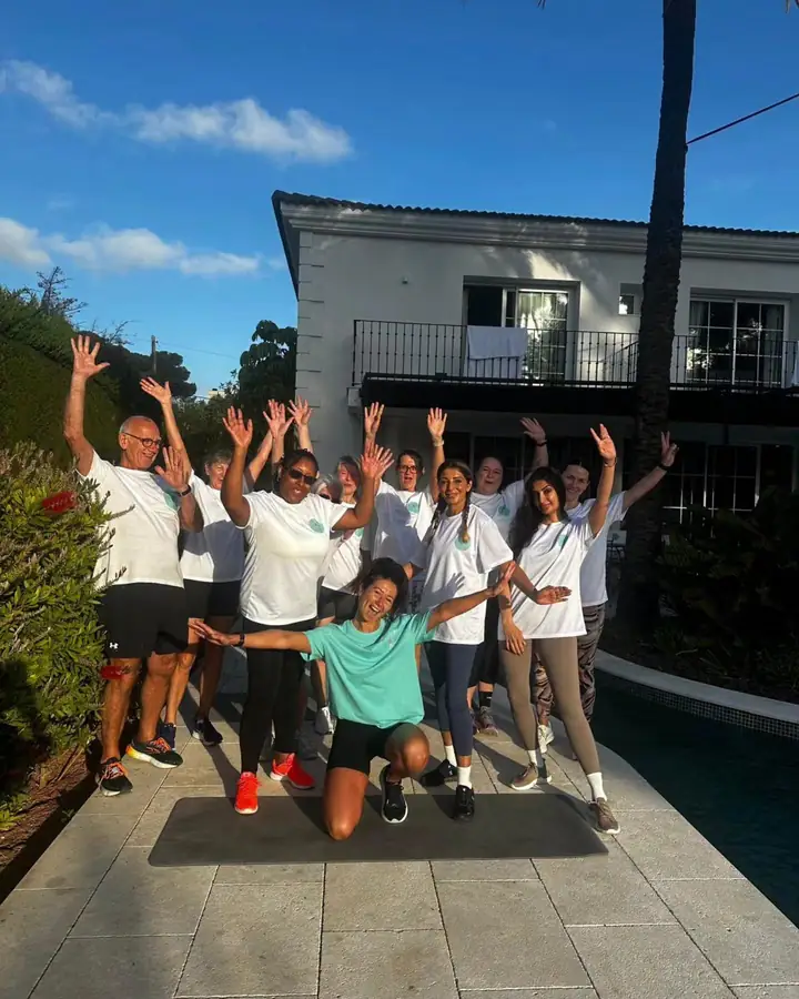 A group in white t-shirts with their arms in the air beside a pool and a white villa, one woman kneeling at the front.