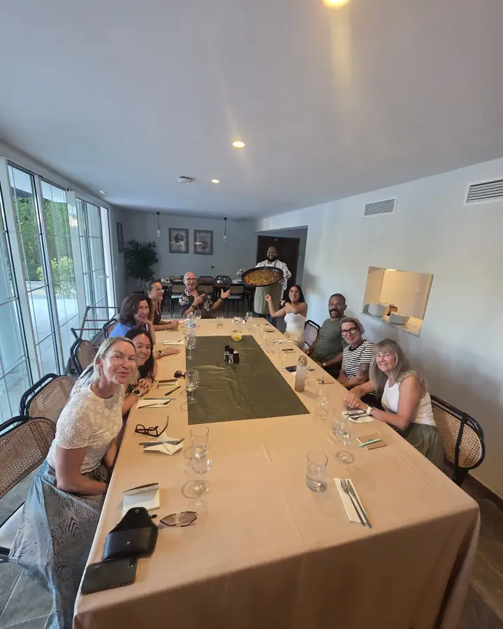 Guests around a long dining table at the villa while the chef serves paella.