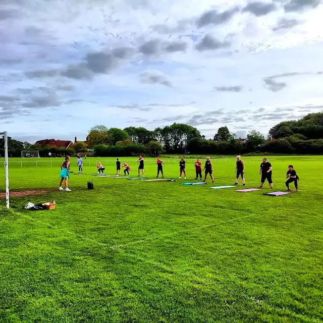 A line of people training on mats across a wide green field under a cloudy sky.