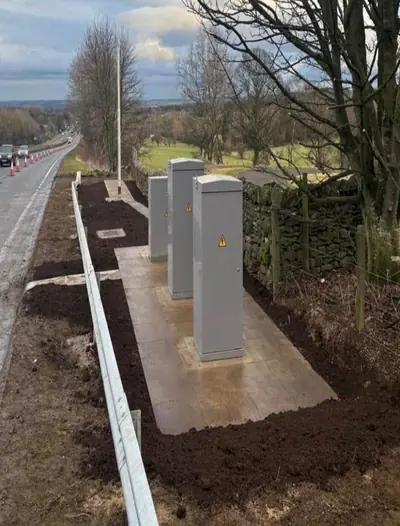 Three grey roadside cabinets standing on a new concrete base beside a rural road, with fresh topsoil dressed along the verge, a drystone wall behind and cones on the carriageway.