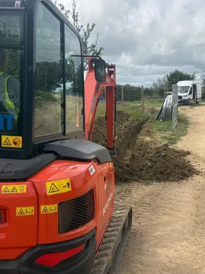 Orange mini excavator parked alongside a shallow trench cut into the verge of a stone farm track, with coils of black ducting stacked further along.