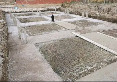 A grid of concrete bases cast between excavated foundation strips, with short concrete columns standing and a brick wall behind.