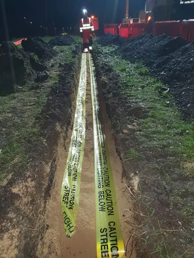 Yellow warning tape printed with caution street lighting below, laid the full length of a night trench through a verge, with an operative in red hi-vis at the far end.