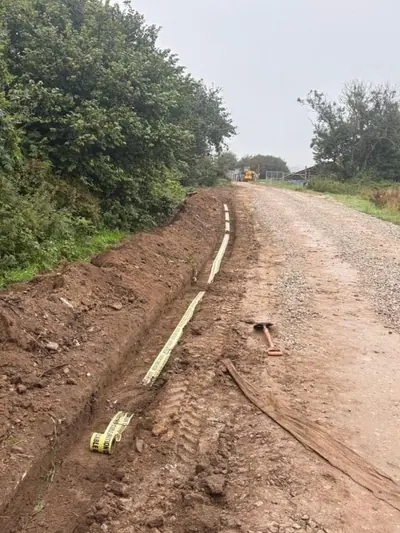 Yellow warning tape laid along the bottom of a shallow trench beside a stone track, with geotextile matting rolled out on the track surface.