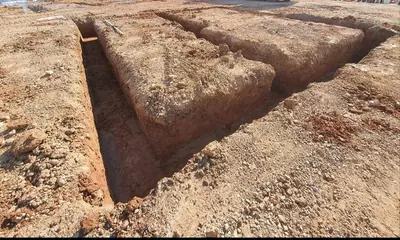 Deep strip foundation trenches cut square into orange clay ground, forming a rectangle around an untouched platform.