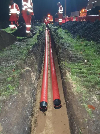 Two red ducts laid side by side in a narrow trench through a grass verge at night, with operatives in red hi-vis and red barriers behind.