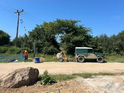 A cable drum trailer and a coil of blue ducting parked on a stone compound in bright sun, with a green Land Rover, barrier fencing and an overhead line behind.
