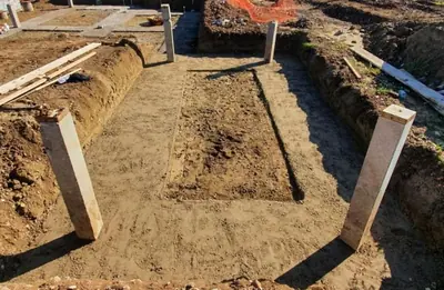 A foundation trench dug around a rectangular platform with concrete posts standing at the corners and timber boards laid across the excavation.