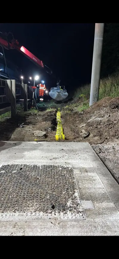 A heavy duty chamber cover set flush in a finished concrete surround at night, with a backfilled trench marked by yellow tape running away towards a lorry and excavator.