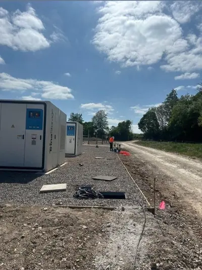 Two containerised battery units on a new stone hardstanding under a blue sky, with cable coiled on the ground beside a pegged and stringed line.