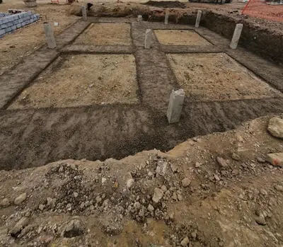An excavated foundation grid with concrete posts standing in it and packs of grey concrete blocks stacked at the edge of the plot.