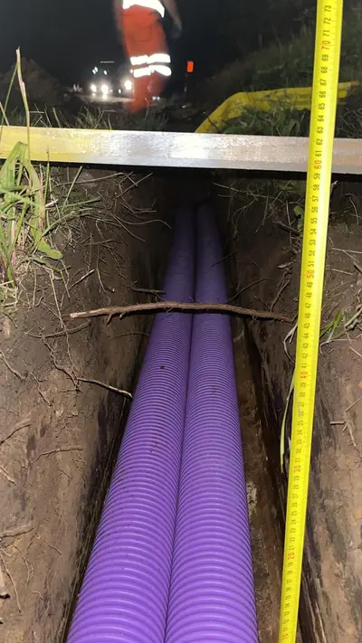 Two purple ducts in the bottom of a trench at night with a tape measure hanging down the trench side and a straight edge laid across the top.