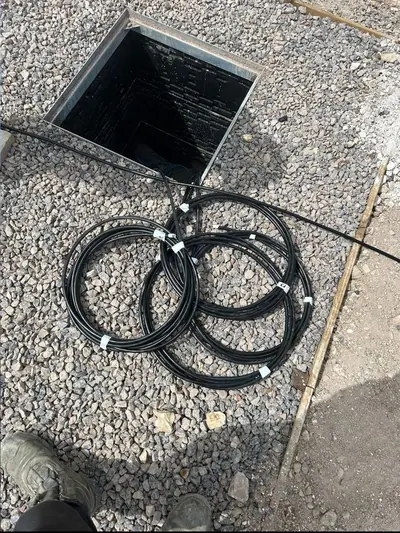 Four coils of black cable with white identification tags laid on stone beside an open square chamber with its frame exposed.