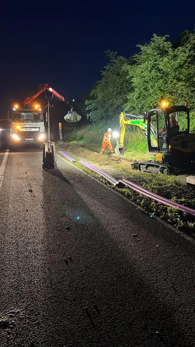 A grab lorry with a red crane and a mini excavator working a verge at night, with purple ducting laid out along the edge of the carriageway and an operative in orange hi-vis between them.