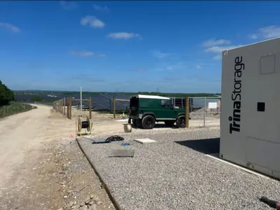 A containerised battery unit at the edge of a stone hardstanding with a timber post fence, a cable drum on the ground and rows of solar panels on the ridge behind.