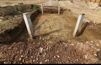 A deeper foundation pit stepped down into red ground with concrete posts standing in the corners and spoil heaped in the foreground.