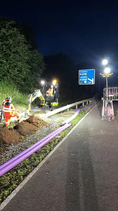 Purple ducting run out along a trunk road verge at night beneath a blue junction sign reading Coventry M69 and Leicester A5460, with a mini excavator, two operatives in orange hi-vis, a lighting tower and lane barriers.