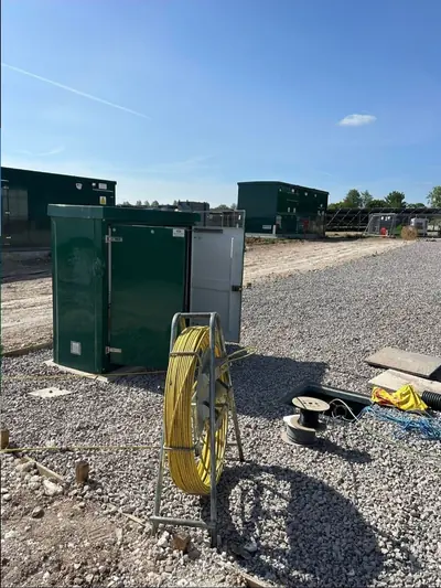 A reel of yellow cable rods on a stand and two cable drums set out on a stone compound beside dark green kiosk enclosures, with rows of solar panels in the distance.