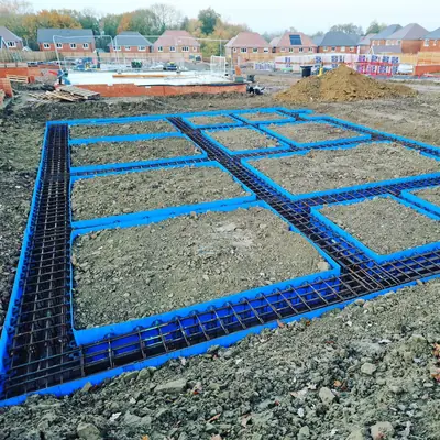 Steel reinforcement mesh and blue edge formwork laid out across a large foundation on a new housing plot, with finished brick houses and scaffolding behind.