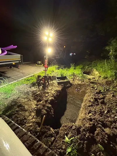A deep excavation opened up in a verge at night under a lighting tower, with a black chamber base and a trailer of purple ducting waiting on the road alongside.
