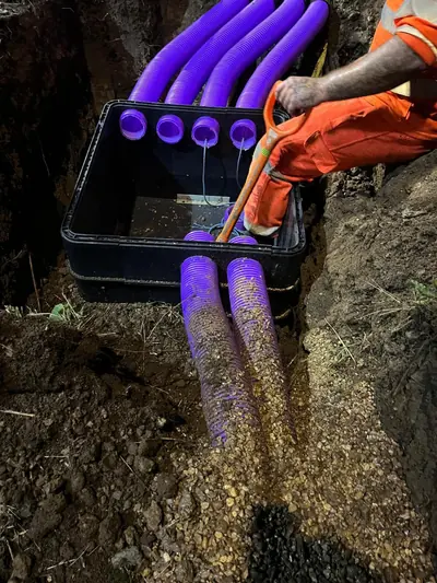 Operative in orange hi-vis kneeling at a black plastic cable box set in a trench, with four purple ducts entering the top and two leaving the front on pea gravel.