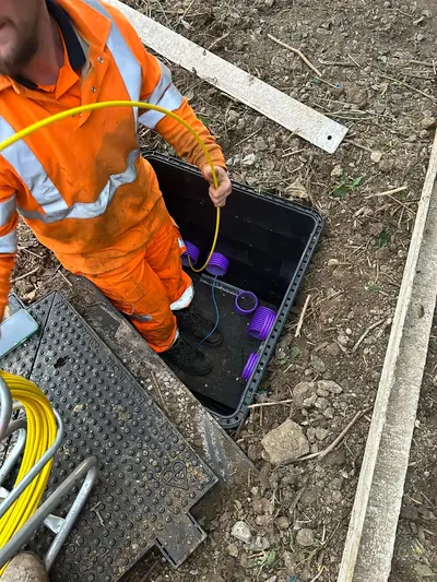 Operative in orange hi-vis standing inside an open cable chamber feeding a yellow fibre cable down into purple duct entries, with the composite chamber cover lifted clear alongside.