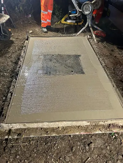 A freshly poured concrete slab being floated smooth around a new chamber cover at night, with timber shuttering still in place and a power float behind.
