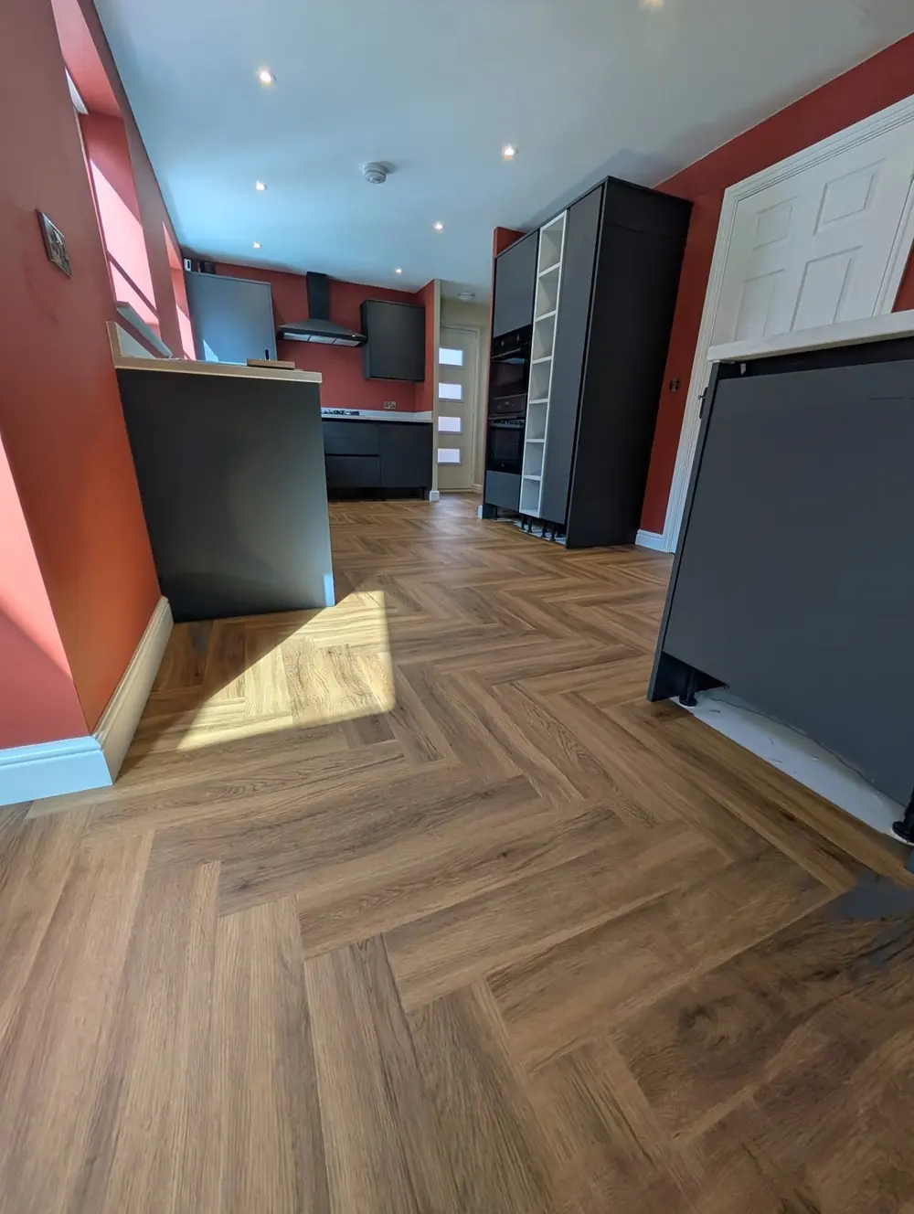 Walnut herringbone LVT in a kitchen diner with a terracotta wall, sunlight falling across the planks.