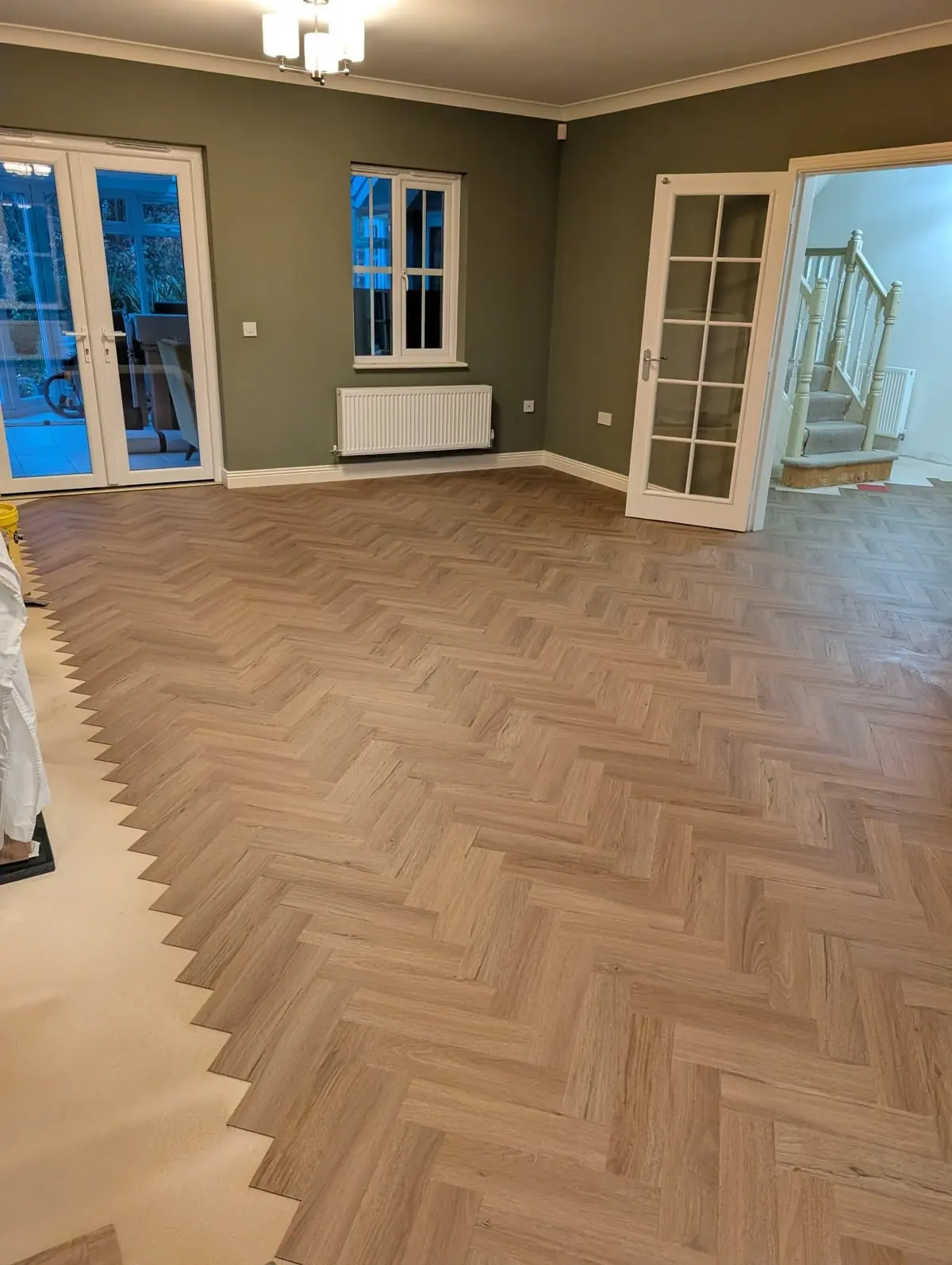 Warm oak herringbone LVT filling an olive green living room, with white French doors and a staircase edge.